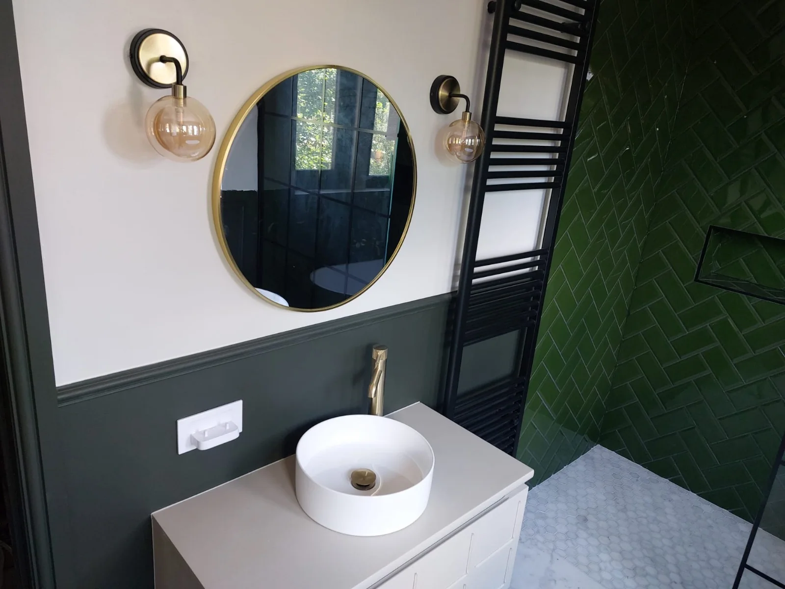 Finished bathroom with round brass mirror, globe wall lights and vessel basin beside a green herringbone shower